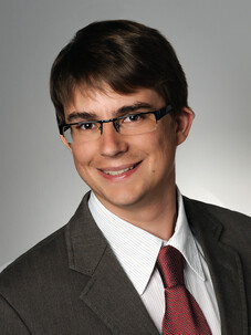 Simon Moser im Anzug mit Brille, vor grauem Verlaufshintergrund.